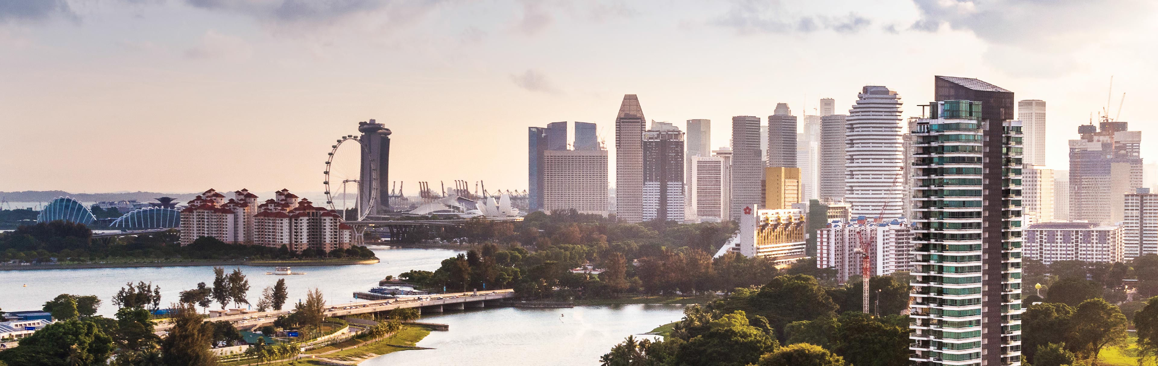 Der Blick auf die Skyline von Singapur zeigt moderne Hochhäuser, die sich entlang der Küste erstrecken. Links ist ein großes Rad zu sehen, während im Hintergrund die Marina Bay Sands und andere markante Gebäude zu erkennen sind. Ein Fluss fließt durch die Szene, umgeben von üppiger Vegetation.