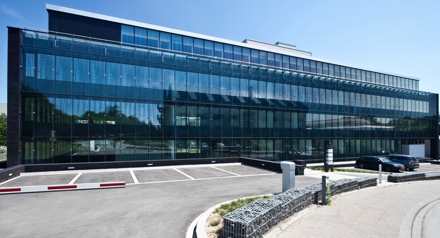 Ein modernes, mehrstöckiges Bürogebäude mit großen Glasfenstern, das von Sonnenlicht erleuchtet wird. Vor dem Gebäude befindet sich ein Parkplatz mit mehreren Autos. Im Hintergrund sind grüne Bäume sichtbar, die das Umfeld auflockern.