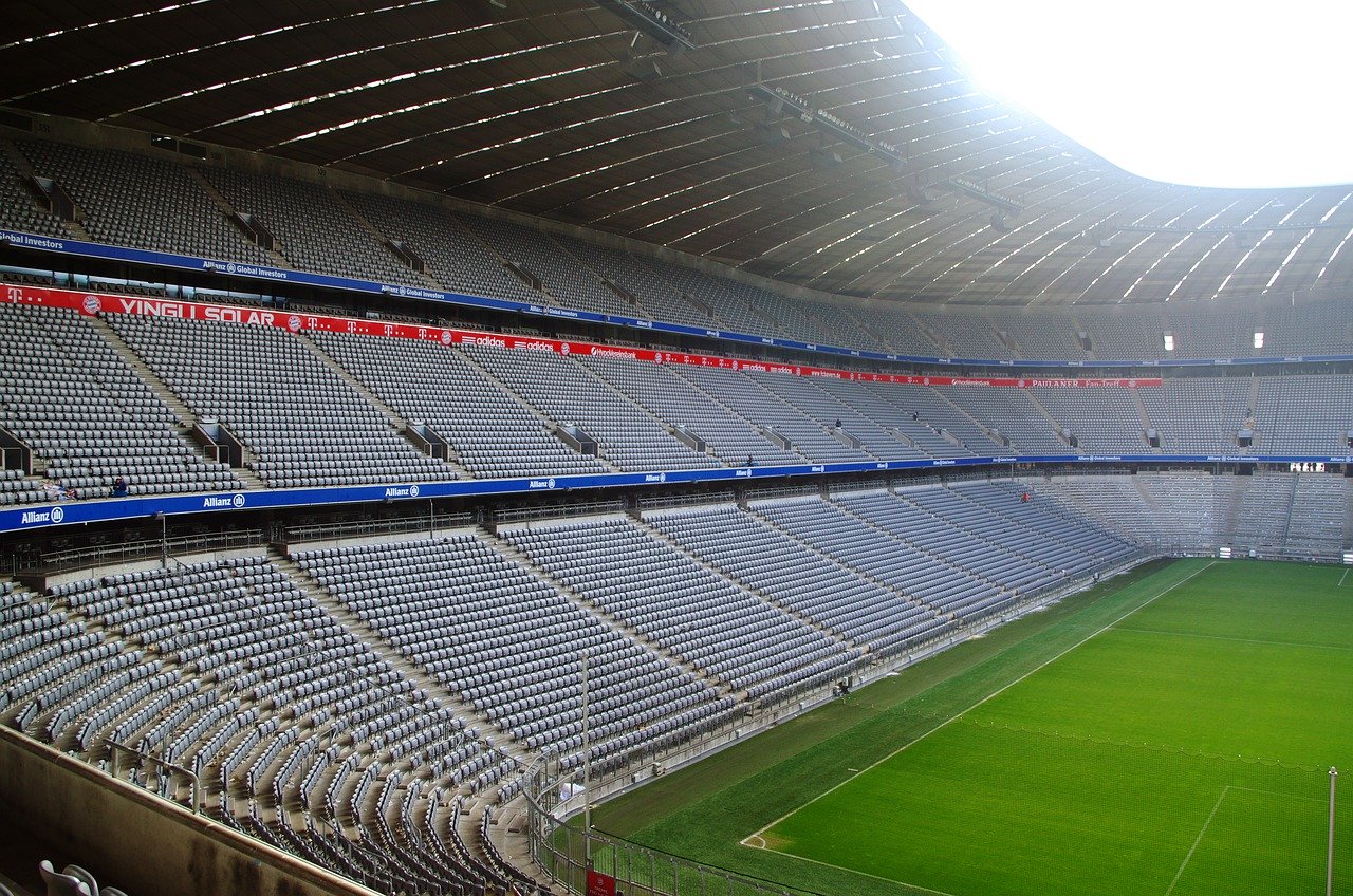 A large, empty stadium is shown, featuring numerous gray seats arranged in rows. The grass pitch is visible in the foreground, surrounded by fencing. A part of the stadium's roof is softly illuminated, indicating daylight. The scene conveys a sense of anticipation for upcoming events.