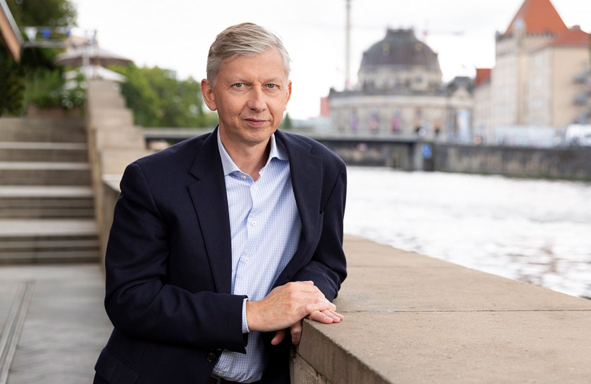 Ein Mann in einem dunklen Blazer und einem hellen Hemd steht an einer Mauer mit Blick auf ein Gewässer. Im Hintergrund sind Gebäude und ein Stadtbild sichtbar.