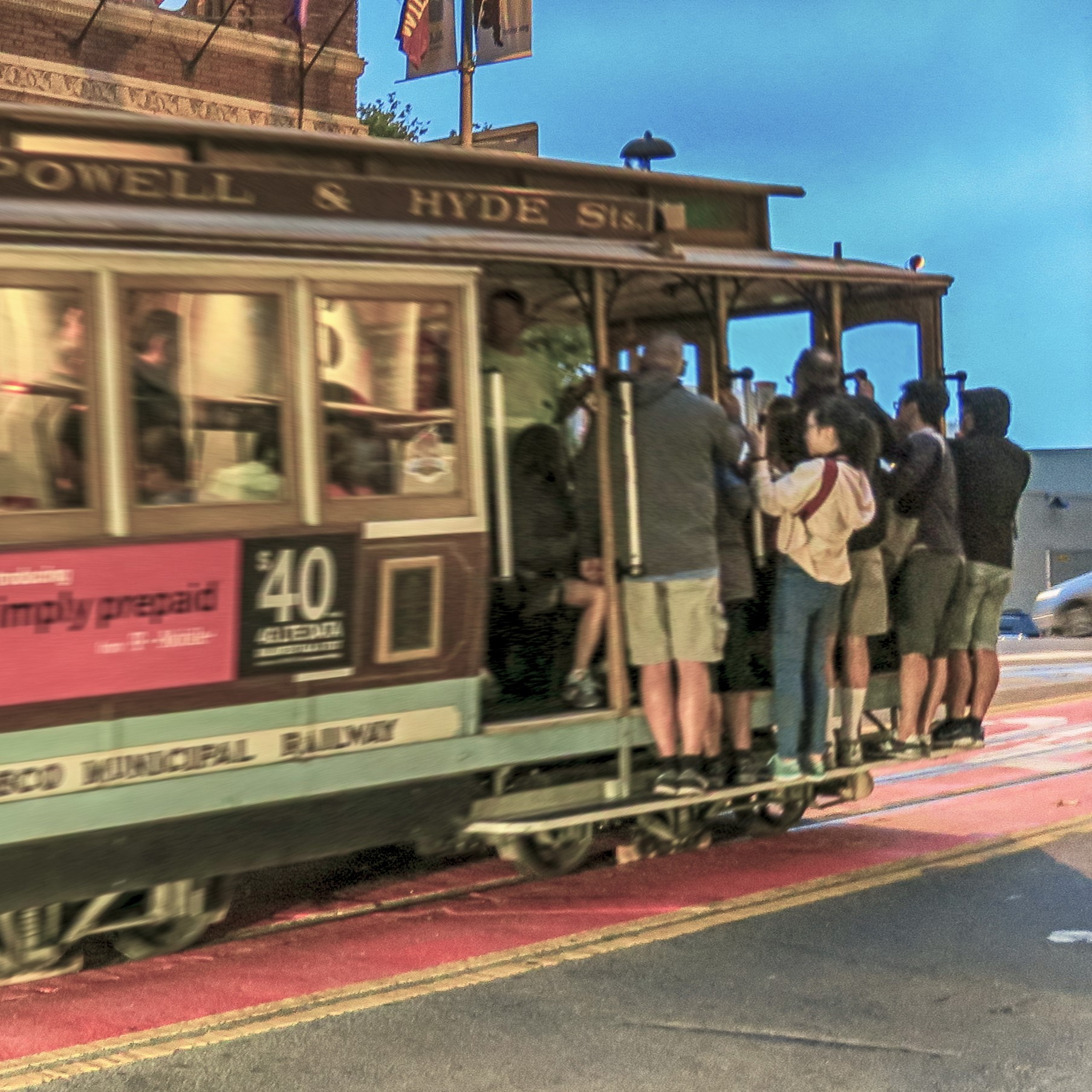 A classic cable car, brightly colored, is filled with numerous passengers standing on the exterior, holding onto poles. The scene captures the bustling atmosphere of a city street, suggesting a lively public transport experience at dusk.