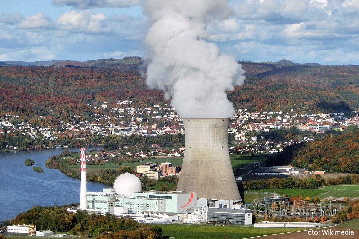 The image shows a nuclear power plant with a prominent cooling tower emitting steam. In the background, a river runs alongside forested hills and a small town with houses. The scene captures an industrial landscape integrated with natural elements.