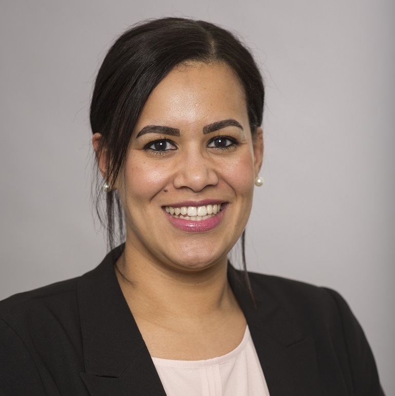 Porträt einer lächelnden Frau mit dunklem Haar und Ohrstöpseln, gekleidet in ein schwarzesjackett über einer hellen Bluse. Der Hintergrund ist neutral und grau.